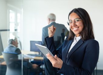 Happy female professional in glasses and suit holding tablet and making winner gesture while two businessmen working behind glass wall. Copy space. Communication concept