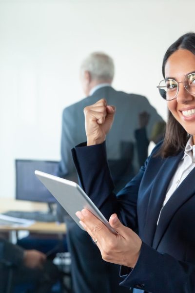 Happy female professional in glasses and suit holding tablet and making winner gesture while two businessmen working behind glass wall. Copy space. Communication concept